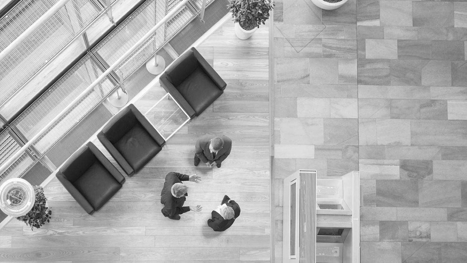 Blick von oben in eine Lobby. Drei Personen in Business-Kleidern begrüssen sich stehend.