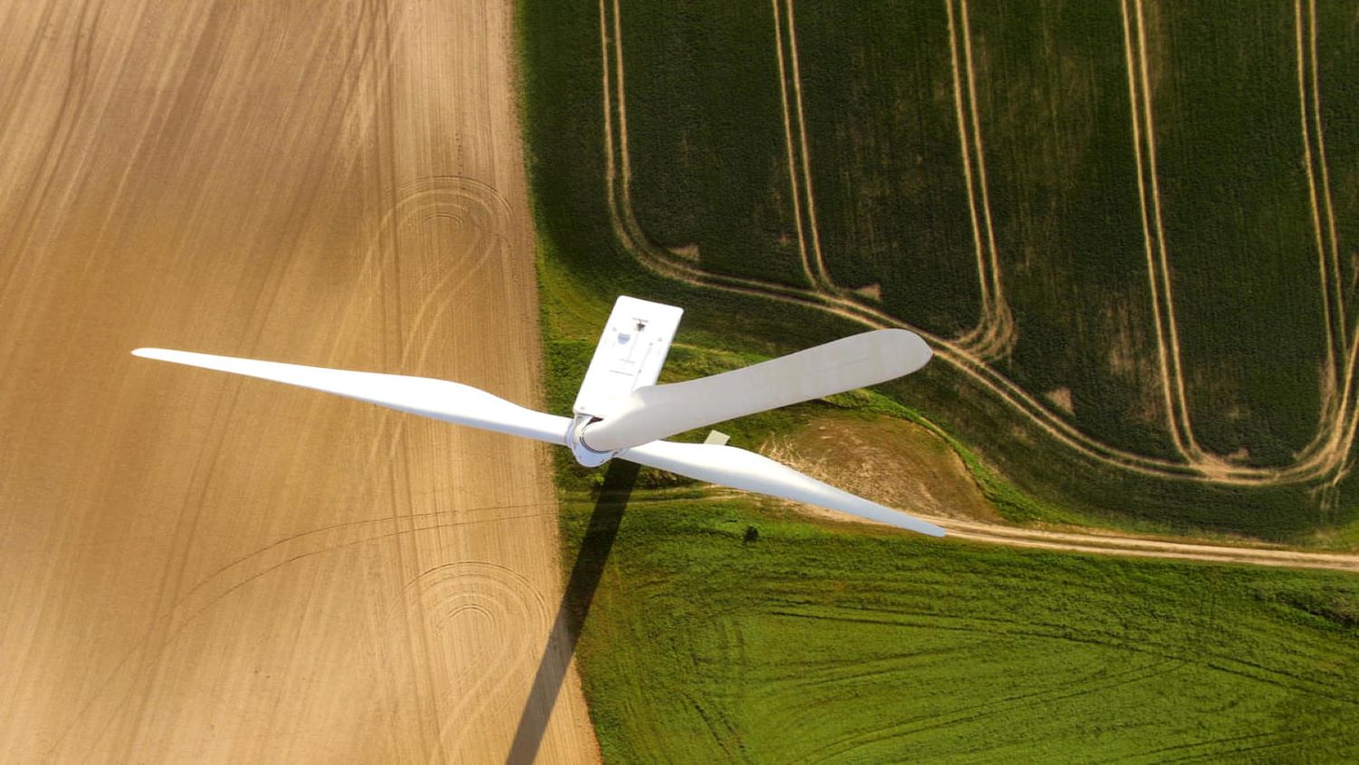 Close-up of the rotor blades of a wind turbine, photographed from above. Far down on the ground, one can see the traces that a tractor has left on an agricultural meadow.