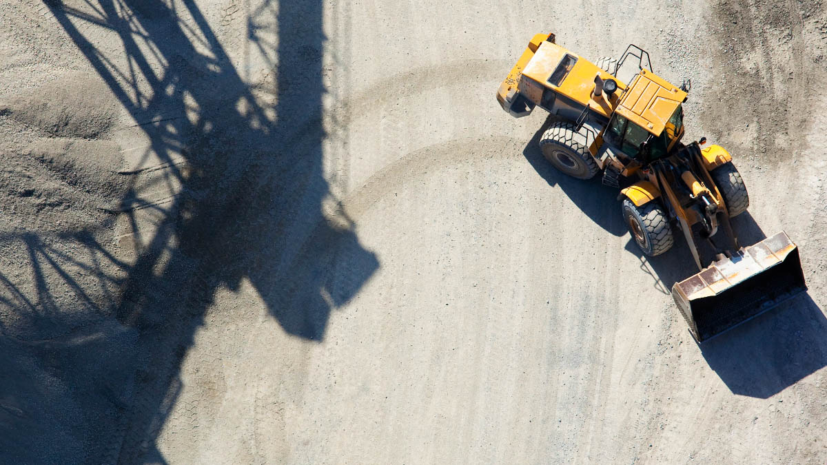 View from above on a heavy excavator, which turns sharply right in a sandy pit. Its tracks cross with the dark shadows of a large industrial plant.