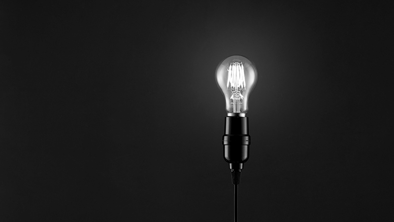 Eine LED-Glühbirne in simpler Fassung mit Stromkabel, fotografiert vor schwarzem Hintergrund. Sie symbolisiert die Wende im Energiezeitalter: hin zu mehr Energieeffizienz und erneuerbaren Energiequellen